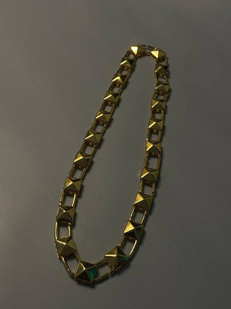 May include: A gold-toned chain necklace featuring a series of repeating pyramid-shaped studs linked together. The metallic links are square-shaped and connected by small rectangular bars, creating a continuous, uniform geometric pattern throughout.