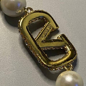 May include: A gold-toned metal letter V pendant featuring embedded clear crystals along the edge, connected to two round white faux pearls. The metal surface displays the text MADE IN ITALY engraved on the side of the letter V component.