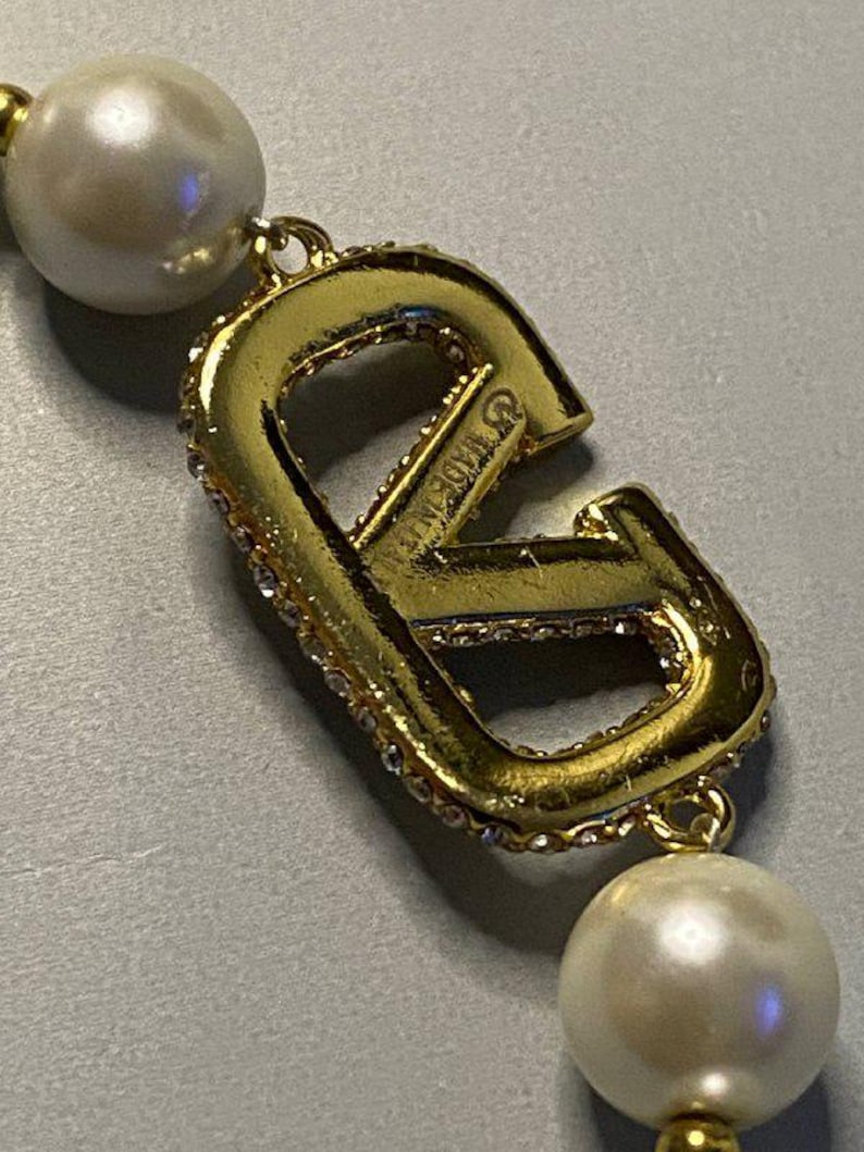 May include: A gold-toned metal letter V pendant featuring embedded clear crystals along the edge, connected to two round white faux pearls. The metal surface displays the text MADE IN ITALY engraved on the side of the letter V component.