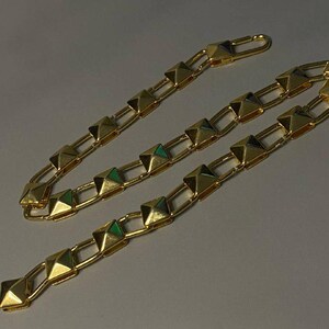 May include: A gold-toned chain necklace featuring a series of repeating pyramid-shaped studs linked together by rectangular metal bars. The chain includes a hook-style clasp mechanism at one end, showcasing a polished metallic finish throughout.