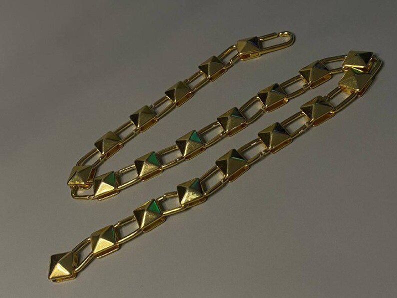 May include: A gold-toned chain necklace featuring a series of repeating pyramid-shaped studs linked together by rectangular metal bars. The chain includes a hook-style clasp mechanism at one end, showcasing a polished metallic finish throughout.