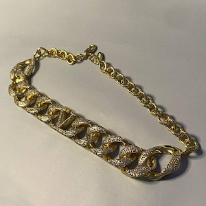 Puede incluir: Un collar de cadena en tono dorado con una serie de eslabones grandes entrelazados. La parte frontal del collar est&aacute; adornada con cristales claros y brillantes incrustados en el metal, mientras que la parte posterior es una cadena sencilla.
