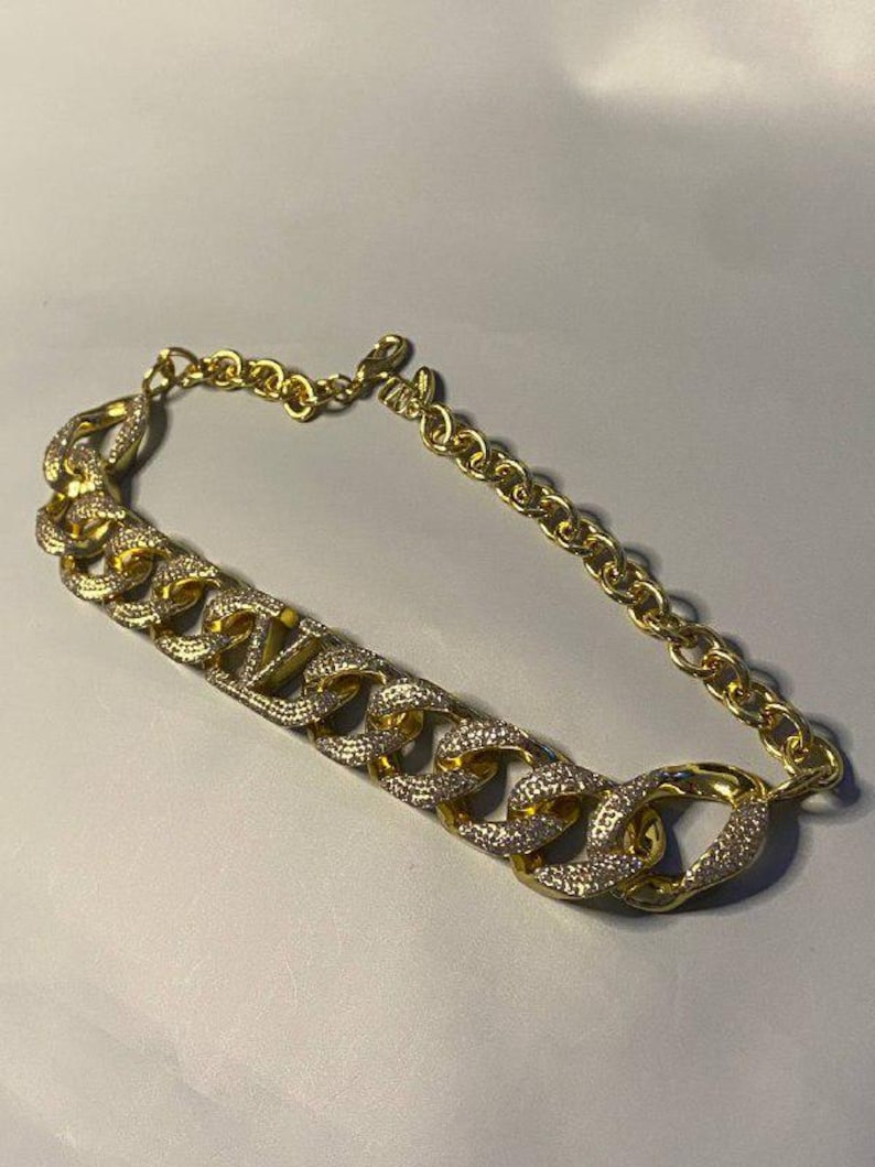 Puede incluir: Un collar de cadena en tono dorado con una serie de eslabones grandes entrelazados. La parte frontal del collar est&aacute; adornada con cristales claros y brillantes incrustados en el metal, mientras que la parte posterior es una cadena sencilla.