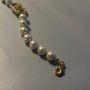 May include: A bracelet featuring a string of white spherical beads separated by small gold-toned spacers. A gold-toned lobster claw clasp is attached to one end. A gold-toned charm encrusted with small clear crystals is integrated into the bead strand.