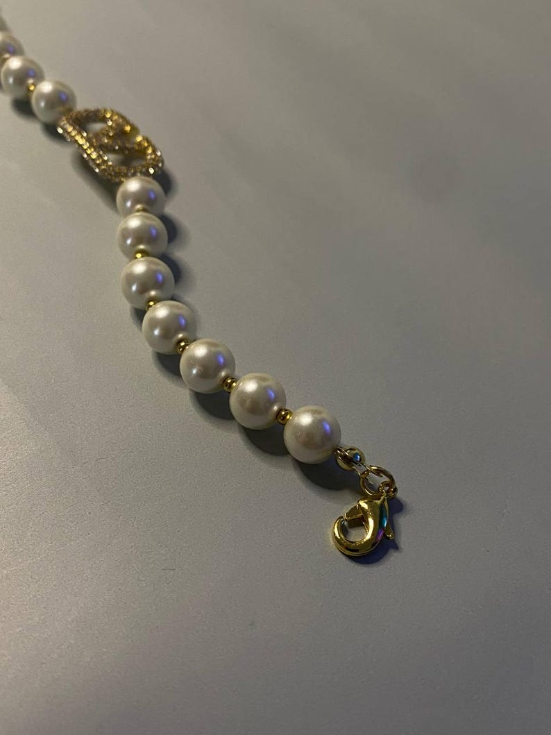 May include: A bracelet featuring a string of white spherical beads separated by small gold-toned spacers. A gold-toned lobster claw clasp is attached to one end. A gold-toned charm encrusted with small clear crystals is integrated into the bead strand.