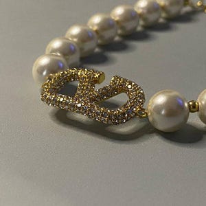 May include: A necklace featuring a strand of round white faux pearls interspersed with small gold-toned beads. The centerpiece is a gold-toned metal letter V logo encrusted with clear sparkling rhinestones, positioned between the pearl segments.