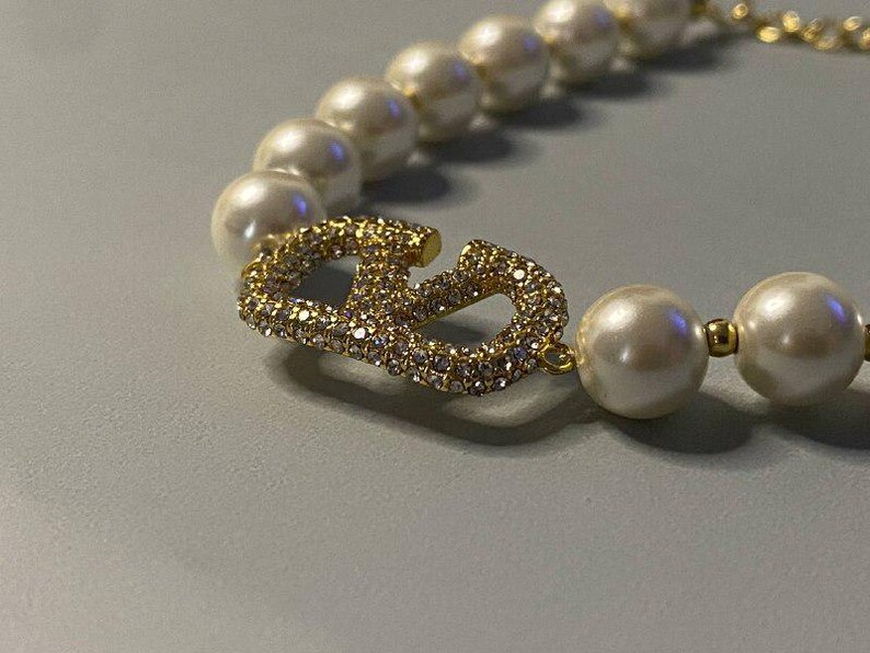 May include: A necklace featuring a strand of round white faux pearls interspersed with small gold-toned beads. The centerpiece is a gold-toned metal letter V logo encrusted with clear sparkling rhinestones, positioned between the pearl segments.
