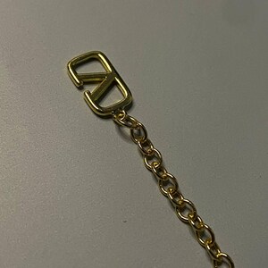 May include: A gold-toned metal chain featuring a prominent V-shaped logo charm at the end. The charm consists of a stylized letter V integrated into a rectangular frame, connected to a series of small, uniform circular links forming a delicate chain.