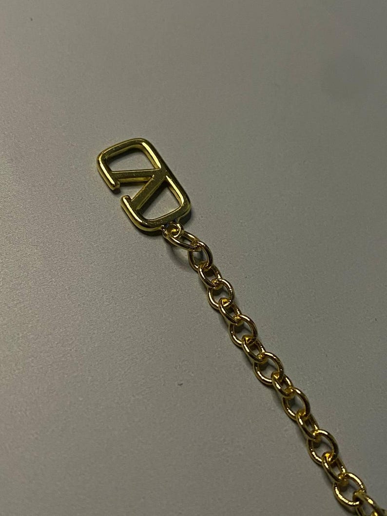 May include: A gold-toned metal chain featuring a prominent V-shaped logo charm at the end. The charm consists of a stylized letter V integrated into a rectangular frame, connected to a series of small, uniform circular links forming a delicate chain.