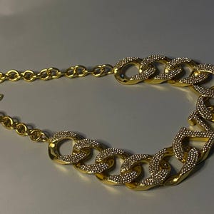 Puede incluir: Un collar de cadena en tono dorado que presenta una serie de eslabones ovalados grandes y entrelazados. La secci&oacute;n frontal del collar est&aacute; adornada con cristales claros y brillantes incrustados en la superficie de los eslabones, mientras que la parte posterior consta de eslabones circulares lisos en tono dorado.