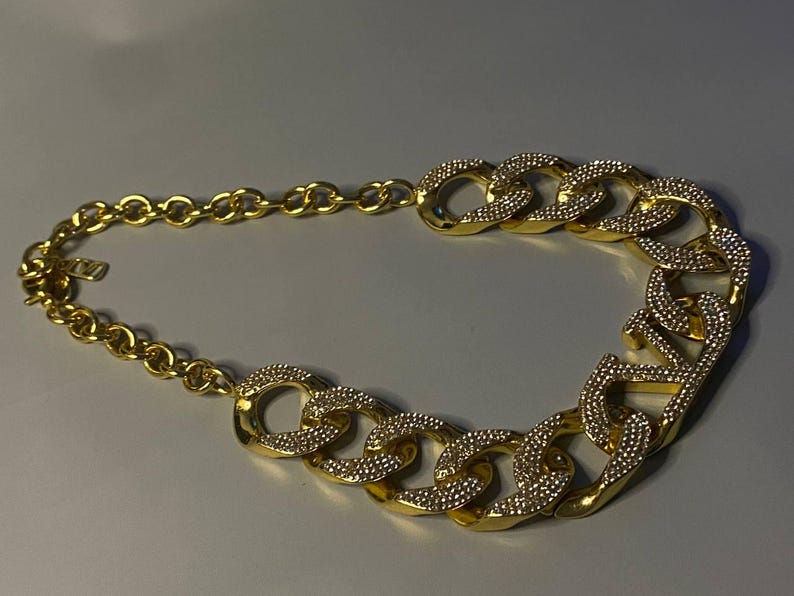 Puede incluir: Un collar de cadena en tono dorado que presenta una serie de eslabones ovalados grandes y entrelazados. La secci&oacute;n frontal del collar est&aacute; adornada con cristales claros y brillantes incrustados en la superficie de los eslabones, mientras que la parte posterior consta de eslabones circulares lisos en tono dorado.
