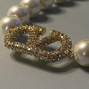 May include: A necklace featuring a series of round white pearls interspersed with a central gold-toned metal V-shaped charm. The charm is encrusted with numerous small, clear, sparkling crystals that catch the light, creating a bright, reflective surface.