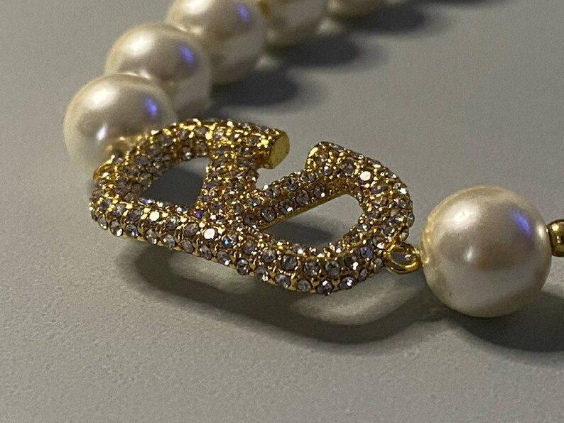 May include: A necklace featuring a series of round white pearls interspersed with a central gold-toned metal V-shaped charm. The charm is encrusted with numerous small, clear, sparkling crystals that catch the light, creating a bright, reflective surface.