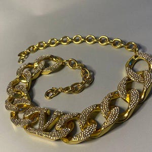 Puede incluir: Un collar de cadena en tono dorado con eslabones grandes, gruesos y entrelazados. Cada eslab&oacute;n est&aacute; parcialmente incrustado con peque&ntilde;os cristales claros y brillantes. El collar incluye una secci&oacute;n de cadena m&aacute;s fina con un cierre de mosquet&oacute;n.