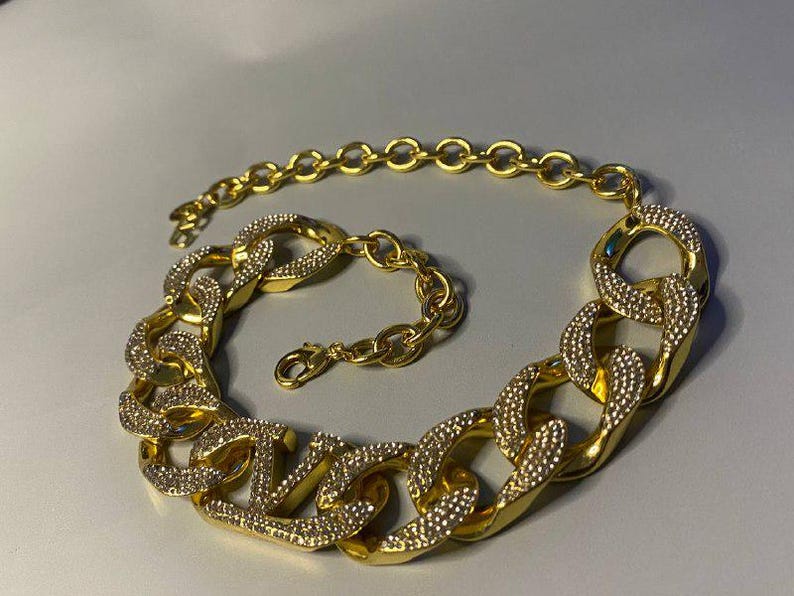 Puede incluir: Un collar de cadena en tono dorado con eslabones grandes, gruesos y entrelazados. Cada eslab&oacute;n est&aacute; parcialmente incrustado con peque&ntilde;os cristales claros y brillantes. El collar incluye una secci&oacute;n de cadena m&aacute;s fina con un cierre de mosquet&oacute;n.