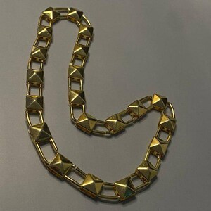 May include: A gold-toned chain necklace featuring a series of repeating square pyramid-shaped studs linked together by thin metal bars. The necklace has a polished, reflective finish and is arranged in a curved, open-ended loop shape.