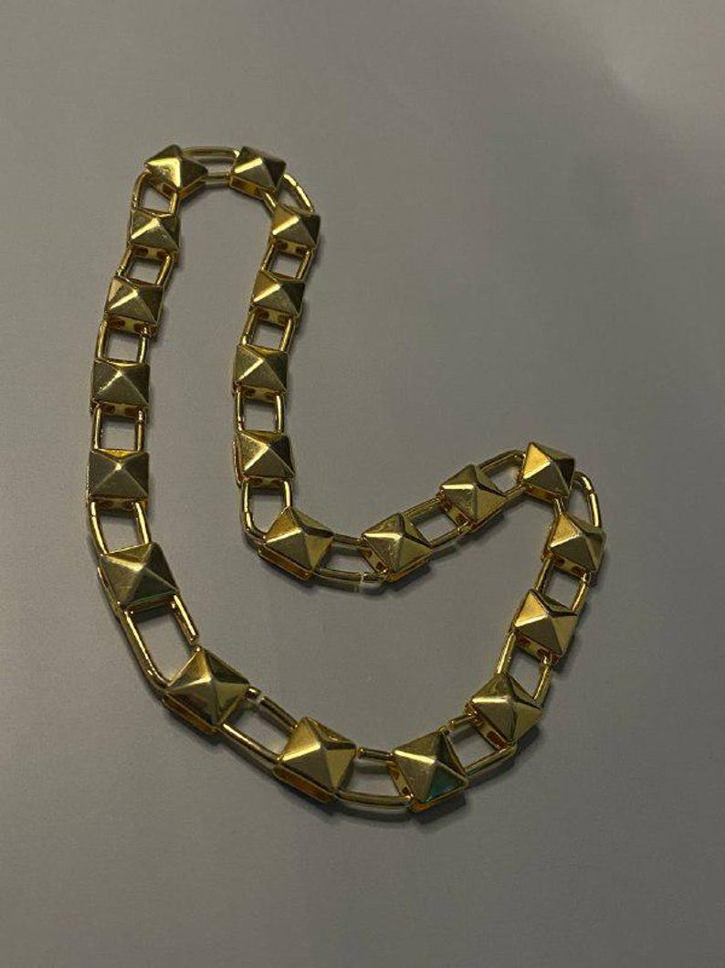 May include: A gold-toned chain necklace featuring a series of repeating square pyramid-shaped studs linked together by thin metal bars. The necklace has a polished, reflective finish and is arranged in a curved, open-ended loop shape.