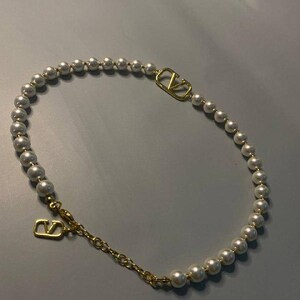May include: A necklace featuring a strand of white spherical pearls interspersed with a gold-toned metal V-shaped logo emblem. The piece includes a gold-toned metal chain extension and a lobster clasp closure with a dangling V-shaped logo charm.
