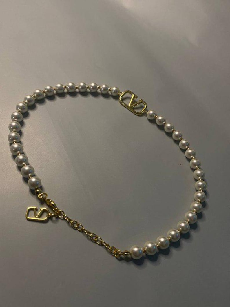 May include: A necklace featuring a strand of white spherical pearls interspersed with a gold-toned metal V-shaped logo emblem. The piece includes a gold-toned metal chain extension and a lobster clasp closure with a dangling V-shaped logo charm.