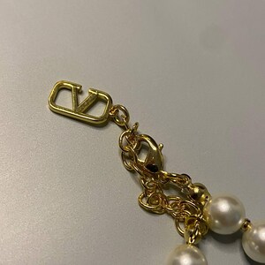 May include: A gold-toned necklace featuring a series of round white pearls and a metallic gold-toned V-logo charm. The necklace includes a gold-toned chain with a lobster claw clasp and a small dangling V-logo emblem at the end of the extender chain.
