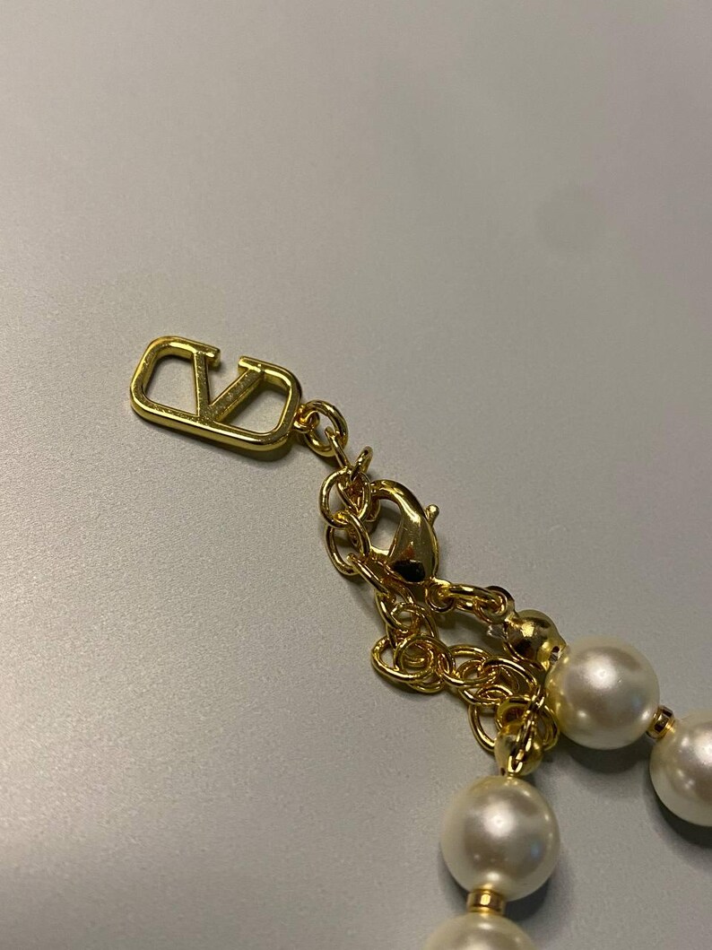 May include: A gold-toned necklace featuring a series of round white pearls and a metallic gold-toned V-logo charm. The necklace includes a gold-toned chain with a lobster claw clasp and a small dangling V-logo emblem at the end of the extender chain.