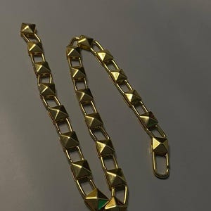 May include: A gold-toned chain featuring a series of repeating pyramid-shaped studs linked together. The chain includes a lobster claw clasp at one end, designed with a polished metallic finish that reflects light across its geometric surfaces.