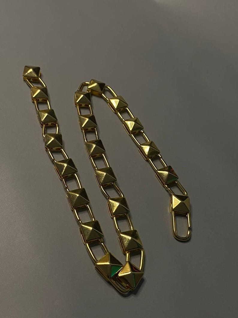 May include: A gold-toned chain featuring a series of repeating pyramid-shaped studs linked together. The chain includes a lobster claw clasp at one end, designed with a polished metallic finish that reflects light across its geometric surfaces.
