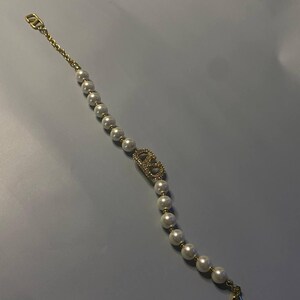 May include: A gold-tone bracelet featuring a series of white spherical faux pearls strung on a delicate chain. The center of the bracelet displays a gold-tone logo emblem encrusted with small clear crystals, finished with a lobster claw clasp.