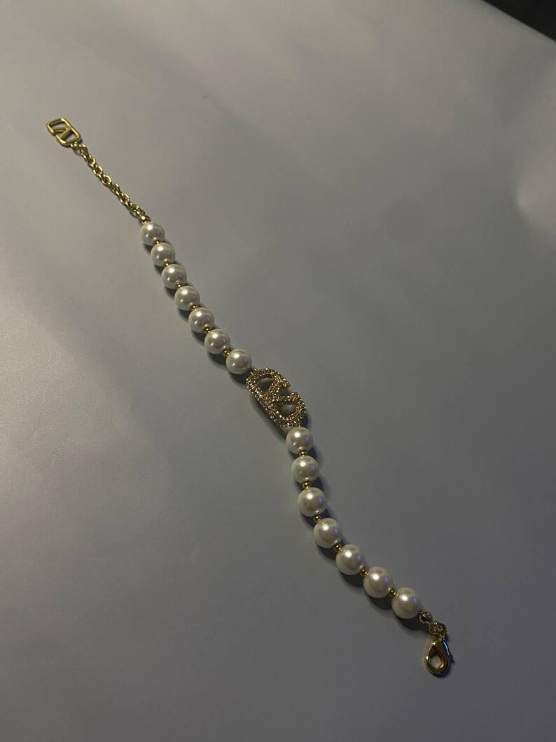 May include: A gold-tone bracelet featuring a series of white spherical faux pearls strung on a delicate chain. The center of the bracelet displays a gold-tone logo emblem encrusted with small clear crystals, finished with a lobster claw clasp.