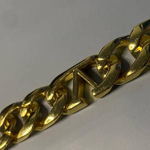 Puede incluir: Un collar de cadena de metal en tono dorado con eslabones gruesos entrelazados. Uno de los eslabones centrales tiene forma de letra V may&uacute;scula, integrada perfectamente en el patr&oacute;n repetitivo de la cadena met&aacute;lica pulida y brillante.