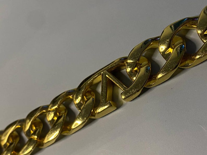 Puede incluir: Un collar de cadena de metal en tono dorado con eslabones gruesos entrelazados. Uno de los eslabones centrales tiene forma de letra V may&uacute;scula, integrada perfectamente en el patr&oacute;n repetitivo de la cadena met&aacute;lica pulida y brillante.