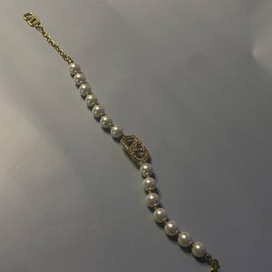 May include: A gold-tone bracelet featuring a series of white spherical faux pearls strung on a delicate chain. The center of the bracelet displays a gold-tone logo emblem encrusted with small clear crystals, finished with a lobster claw clasp.