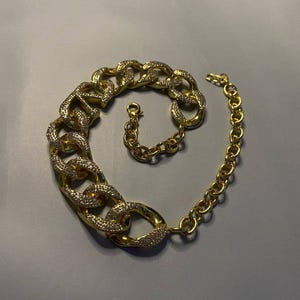 Puede incluir: Un collar de tono dorado que presenta un segmento de cadena grueso y robusto incrustado con peque&ntilde;as piedras transparentes brillantes en un lado, que transiciona hacia una cadena m&aacute;s fina de eslabones circulares lisos con cierre de mosquet&oacute;n.