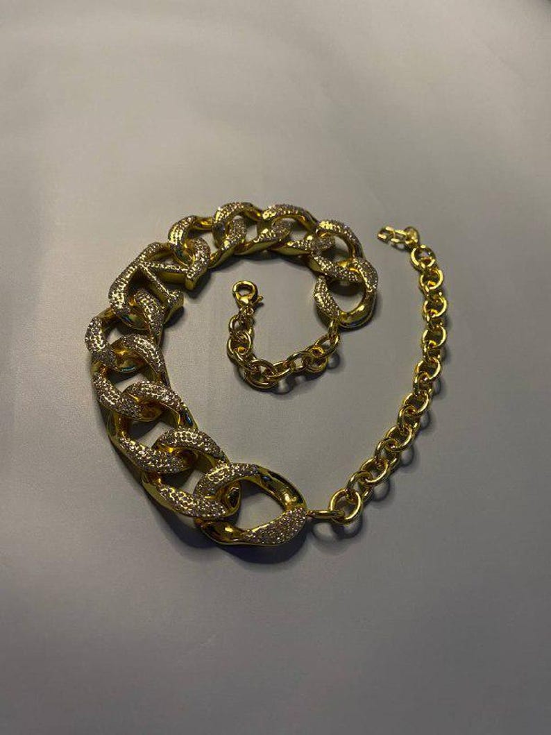 Puede incluir: Un collar de tono dorado que presenta un segmento de cadena grueso y robusto incrustado con peque&ntilde;as piedras transparentes brillantes en un lado, que transiciona hacia una cadena m&aacute;s fina de eslabones circulares lisos con cierre de mosquet&oacute;n.