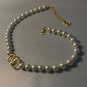 May include: A necklace featuring a strand of white spherical beads interspersed with small gold-toned spacers. A gold-toned V-shaped metal charm is integrated into the strand, and the necklace includes a gold-toned chain extender with a V-shaped tag.