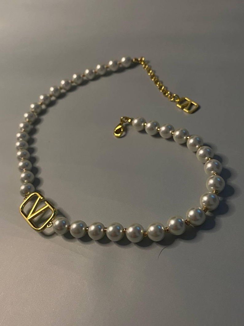 May include: A necklace featuring a strand of white spherical beads interspersed with small gold-toned spacers. A gold-toned V-shaped metal charm is integrated into the strand, and the necklace includes a gold-toned chain extender with a V-shaped tag.