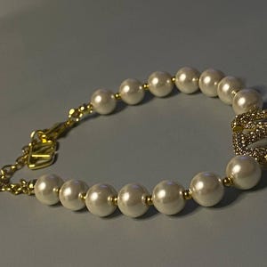 May include: A bracelet featuring a series of white spherical faux pearls separated by small gold-toned metal beads. The bracelet includes a gold-toned chain extension with a clasp and a central gold-toned square charm encrusted with clear crystals.