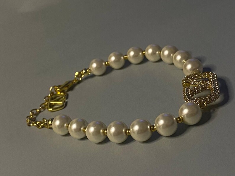 May include: A bracelet featuring a series of white spherical faux pearls separated by small gold-toned metal beads. The bracelet includes a gold-toned chain extension with a clasp and a central gold-toned square charm encrusted with clear crystals.