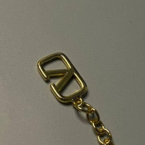 May include: A gold-toned metal charm featuring a stylized V logo, shaped like a V overlapping an inverted V, attached to a delicate gold-toned link chain. The charm has a polished, reflective surface and a smooth, rounded edge design.