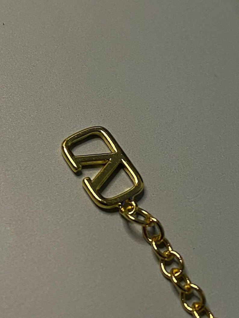 May include: A gold-toned metal charm featuring a stylized V logo, shaped like a V overlapping an inverted V, attached to a delicate gold-toned link chain. The charm has a polished, reflective surface and a smooth, rounded edge design.