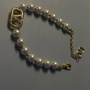 May include: A bracelet featuring a series of white spherical faux pearls separated by small gold-toned beads. The design includes a prominent gold-toned logo charm with a textured surface and a gold-toned chain extension with a small logo pendant.