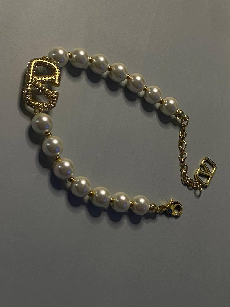 May include: A bracelet featuring a series of white spherical faux pearls separated by small gold-toned beads. The design includes a prominent gold-toned logo charm with a textured surface and a gold-toned chain extension with a small logo pendant.
