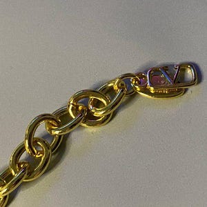 Puede incluir: Un collar de cadena dorada con eslabones ovalados gruesos y entrelazados. El extremo de la cadena termina en una etiqueta de metal dorado pulido que muestra las letras V y L en un dise&ntilde;o de monograma estilizado y superpuesto.