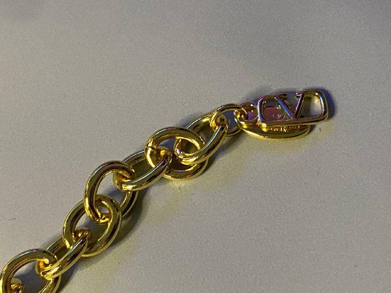 Puede incluir: Un collar de cadena dorada con eslabones ovalados gruesos y entrelazados. El extremo de la cadena termina en una etiqueta de metal dorado pulido que muestra las letras V y L en un dise&ntilde;o de monograma estilizado y superpuesto.