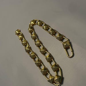 May include: A gold-toned chain featuring a series of repeating pyramid-shaped studs linked together. The chain includes a lobster claw clasp at one end, designed for secure attachment. The metallic finish has a reflective surface throughout.
