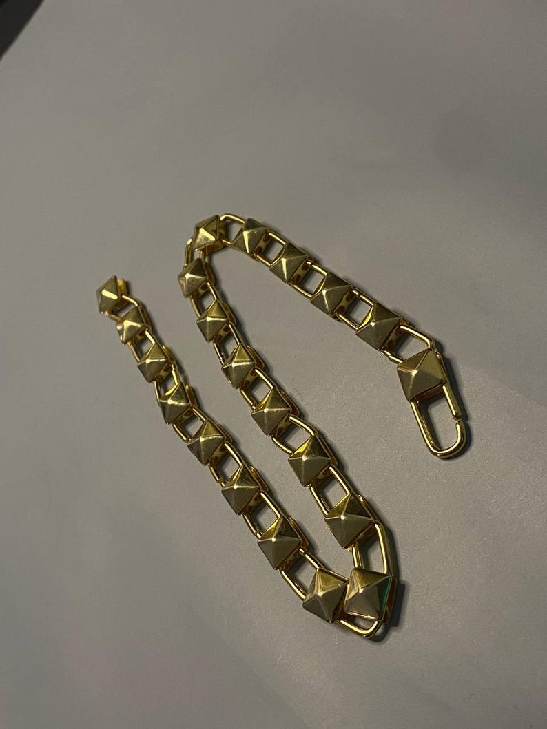 May include: A gold-toned chain featuring a series of repeating pyramid-shaped studs linked together. The chain includes a lobster claw clasp at one end, designed for secure attachment. The metallic finish has a reflective surface throughout.