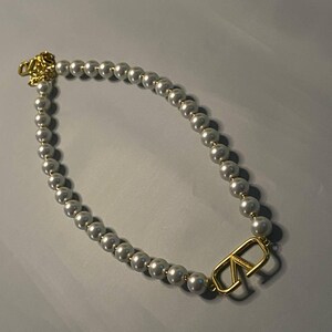 May include: A necklace featuring a strand of white spherical beads with a gold-toned metal V-shaped logo charm integrated into the design. The necklace includes a gold-toned metal clasp mechanism at the end, showcasing a polished finish throughout.