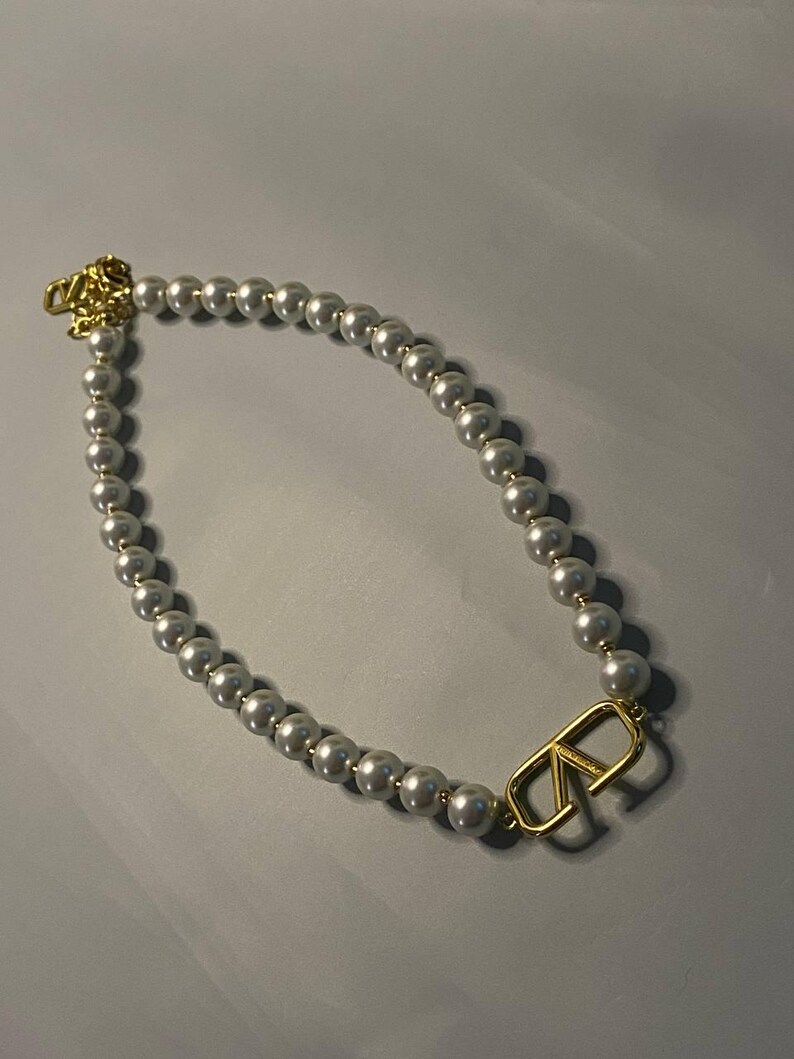 May include: A necklace featuring a strand of white spherical beads with a gold-toned metal V-shaped logo charm integrated into the design. The necklace includes a gold-toned metal clasp mechanism at the end, showcasing a polished finish throughout.