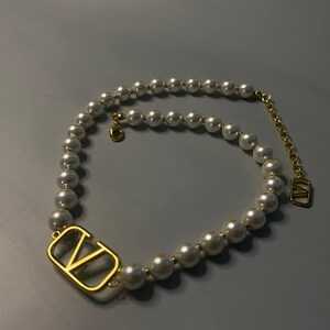 May include: A necklace featuring a strand of white spherical pearls interspersed with small gold-toned beads. The piece includes a prominent gold-toned V-shaped metal emblem and a gold-toned chain extender with a small V-shaped charm at the end.