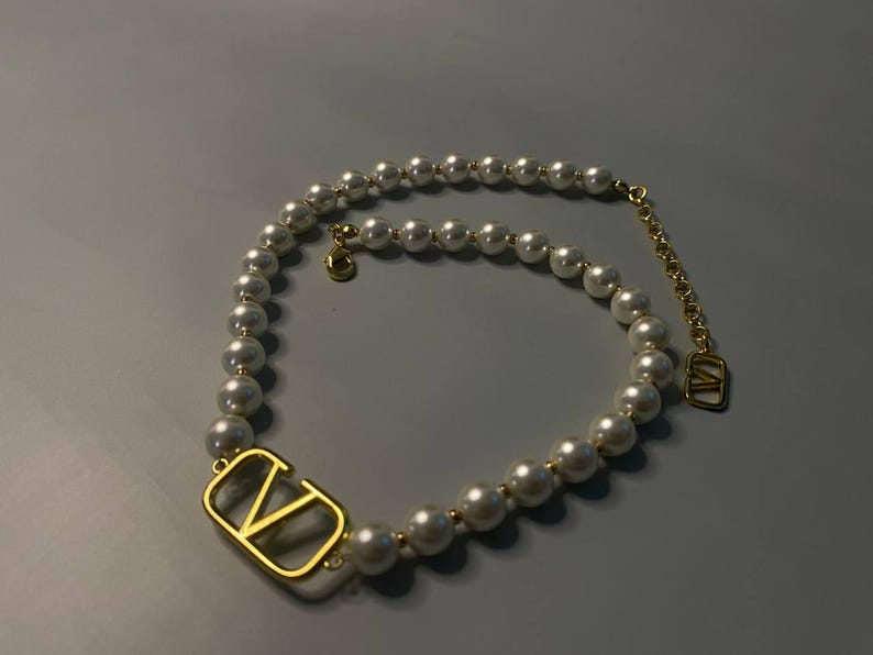 May include: A necklace featuring a strand of white spherical pearls interspersed with small gold-toned beads. The piece includes a prominent gold-toned V-shaped metal emblem and a gold-toned chain extender with a small V-shaped charm at the end.
